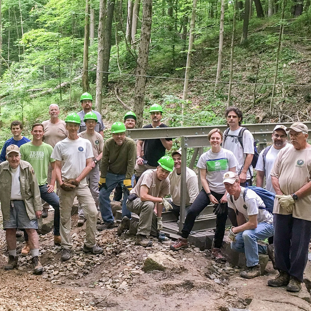 A group of 17 volunteers from Cuyahoga Valley Trails Council, some wearing hard hats, stand in front of a new bridge they are building for a trail in Cuyahoga Valley National Park.